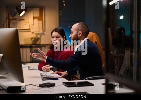 Side view portrait of bearded software developer advising young colleague and pointing at computer screen while collaborating in office Stock Photo