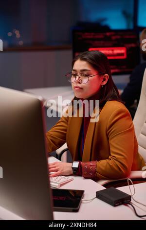 Vertical portrait of young female expert using computer in cybersecurity department with red neon lights Stock Photo
