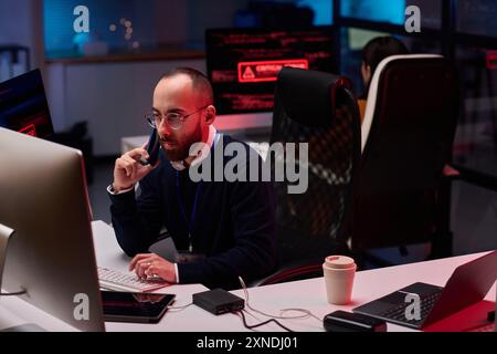 High angle view at bearded adult man using computer with multiple monitors in cybersecurity department and speaking on phone Stock Photo