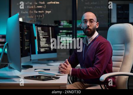 Portrait of bearded network security engineer looking at camera sitting at workplace with code screens in blue light copy space Stock Photo