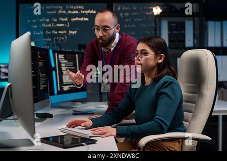 Portrait of young woman using computer in software development office with senior mentor giving advice and pointing at screen copy space Stock Photo