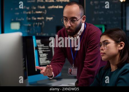 Portrait of bearded IT developer pointing at computer screen while advising young trainee in office with code lines in background Stock Photo