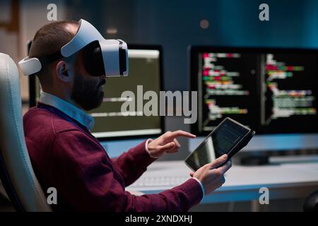 Side view portrait of bearded man wearing VR headset and using tablet working on mixed reality project in blue lights copy space Stock Photo