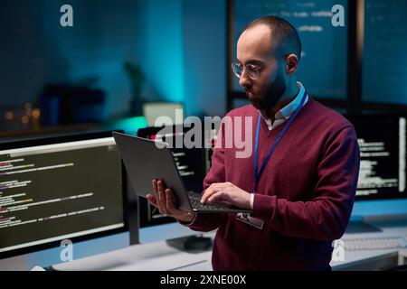 Waist up portrait of bearded adult man holding laptop while standing in cybersecurity office with code lines on screens copy space Stock Photo