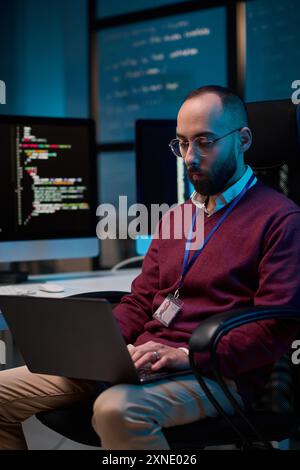 Vertical portrait of adult man using laptop sitting at office workplace in cybersecurity department with code lines and blue neon light Stock Photo