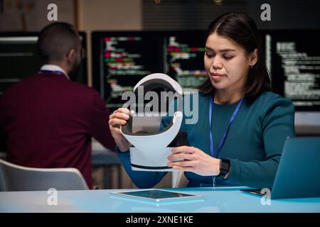 Portrait of young woman setting up VR headset while developing new technology in office with code screens in background, copy space Stock Photo