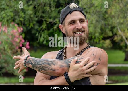 In the foreground, a young instructor smiles as he stretches his arms before beginning his outdoor class. His positive attitude and focus on wellness Stock Photo