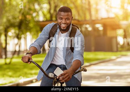 Handsome Black Man With Backpack And Smartphone Riding Bicycle, Posing Outdoors Stock Photo
