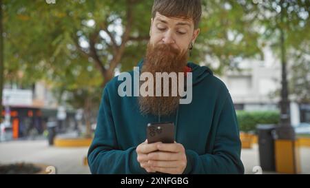 Redheaded bearded young man uses smartphone outdoors in a city park. Stock Photo