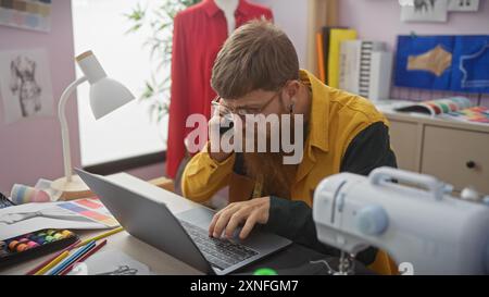 Bearded man using laptop and phone in a tailor's atelier with sewing equipment and colorful fabrics Stock Photo