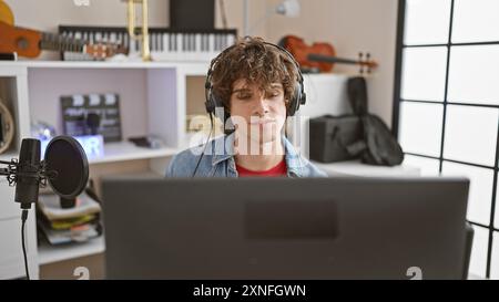 A curly-haired young man wearing headphones sits in a music studio with a microphone and a laptop, concentrated on audio production. Stock Photo