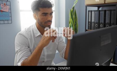 Hispanic man working intently on a computer in a modern office setting, surrounded by files, plants, and colorful sticky notes on a bright day Stock Photo