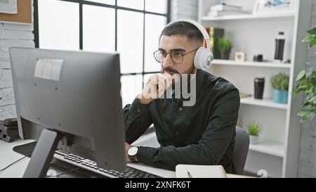 Handsome bearded man with headphones working in a modern office setup, gazing at computer monitor intently. Stock Photo
