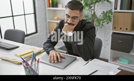 Pensive bearded man in glasses contemplating at desk with laptop in a modern office setting Stock Photo