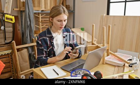 A focused blonde woman using a smartphone in a cluttered carpentry workshop with wood, tools, and a laptop. Stock Photo