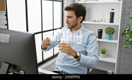 A focused young hispanic man in an office, wearing glasses, a blue shirt, and a watch, interacts with a computer. Stock Photo