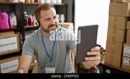 Mature hispanic man with headset using tablet in warehouse full of donations and supplies. Stock Photo