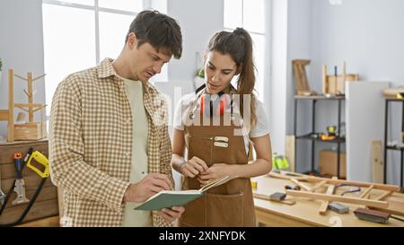 Man and woman, likely carpenters, collaborate in a workshop, examining plans surrounded by woodwork tools. Stock Photo