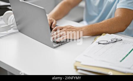 Young hispanic man with a beard working on laptop in modern indoor setting, glasses and documents on table. Stock Photo