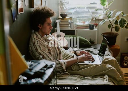 University student writing codes while chilling on soft single bed in his dorm room Stock Photo