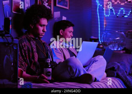 Focused roomies sitting on single bed and drinking beer while working on joint IT project Stock Photo