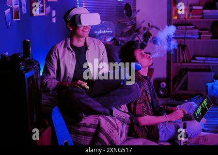 Young man sitting on bed and playing game via VR headset while his friend sitting on floor and vaping while coding with music in headphones Stock Photo
