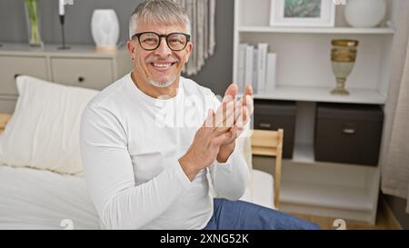 A cheerful mature man with grey hair clapping hands in a modern bedroom interior. Stock Photo