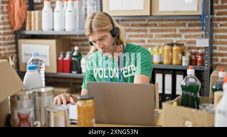 Blond man wearing a headset volunteers at an indoor warehouse, working on a laptop with shelves of supplies in the background. Stock Photo