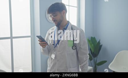 Bearded man in lab coat browsing smartphone in bright hospital room. Stock Photo