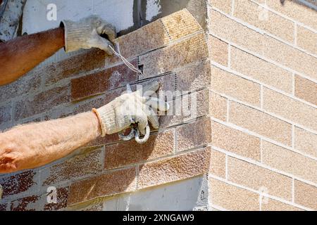 Outdoor facade faux brick wall panel, construction worker makes hole for electrical wiring. Stock Photo
