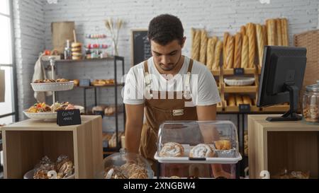 Young hispanic man working in a bakery shop indoors surrounded by various pastries and bread, wearing a white shirt and brown apron concentrating on t Stock Photo