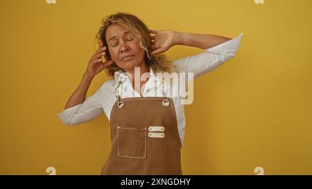 Woman in apron standing against isolated yellow background with eyes closed and hands touching ears representing stress or noise Stock Photo