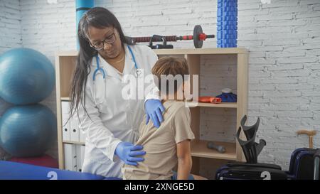 A woman doctor examines a boy in a physiotherapy clinic room, surrounded by rehabilitation equipment. Stock Photo