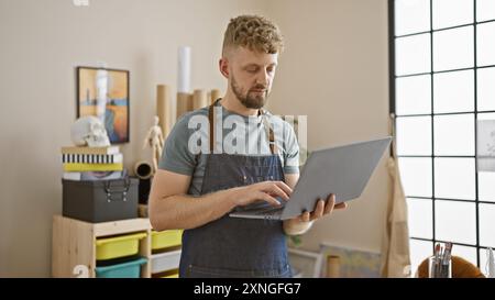 Bearded young caucasian man in studio, wearing apron, using laptop, artist, creative, interior Stock Photo