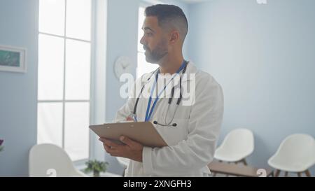 Handsome hispanic man in white coat with stethoscope taking notes in clinic room Stock Photo