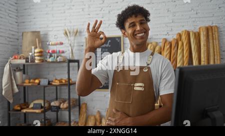 Young man working in a bakery, smiling and giving an ok gesture while standing in front of shelves filled with various types of bread and pastries Stock Photo