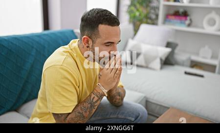 Pensive hispanic man with beard and tattoos sitting indoors wearing yellow shirt Stock Photo