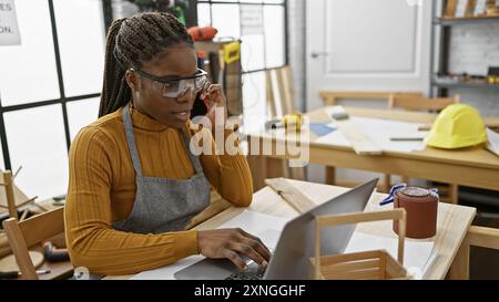 African american woman with braids wearing glasses and using laptop in a woodworking workshop while talking on phone. Stock Photo