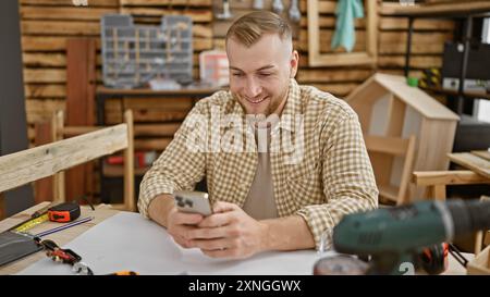 A cheerful young man using smartphone in a cozy carpentry workshop surrounded by tools. Stock Photo