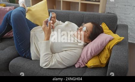 Smiling mature hispanic woman lounging on a sofa at home looking at smartphone. Stock Photo