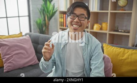 A cheerful asian man wearing glasses sitting comfortably at home on a gray couch surrounded by colorful cushions. Stock Photo