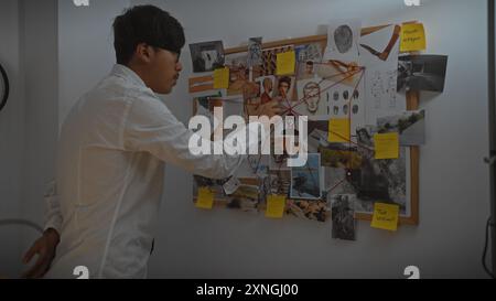 Young asian man examines evidence board in a dimly lit detective's office, embodying focus and analytical skills. Stock Photo