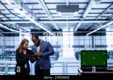 Software developers using green screen laptop in data center, making sure rigs are functioning optimally. Experts in server room using chroma key laptop to monitor energy consumption Stock Photo