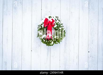 Green pine tree Christmas wreath with red ribbon bow and snow against weathered white-washed wide-plank wood barn door. Vignette and copy space. Stock Photo