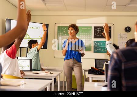In high school, teacher holding tablet and engaging students raising hands in classroom Stock Photo