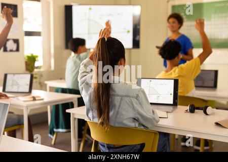 In high school, students raising hands and using laptops during classroom lesson Stock Photo