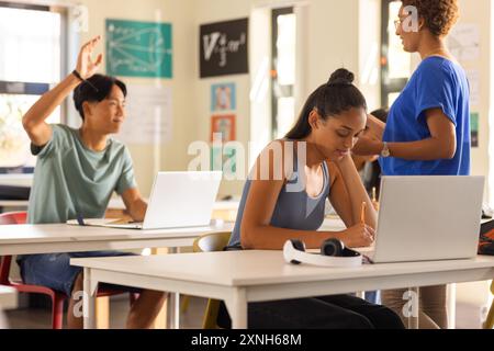 In high school, students using laptops and writing in classroom, teacher assisting Stock Photo