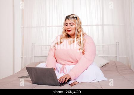 Plus size woman working from home, sitting comfortably on her bed and typing on her laptop Stock Photo