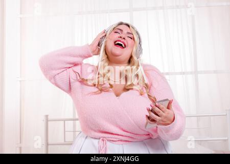Curvy woman is wearing headphones and holding a smartphone while happily singing along to music Stock Photo