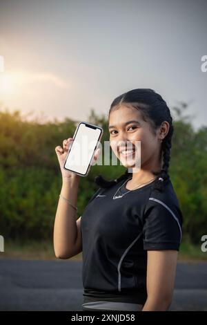 Portrait of sporty young woman in park, she shows her smart phone posing in front of camera in black sportswear, sunset optical flare Stock Photo